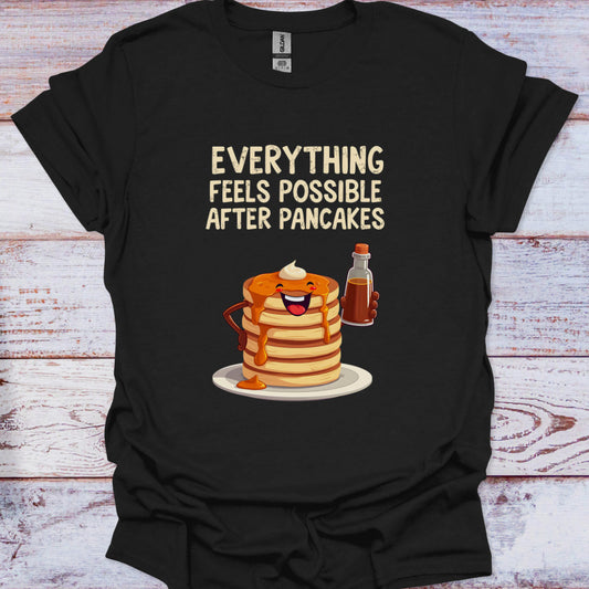 Black t-shirt with a graphic of pancakes and syrup with text 'Everything feels possible after pancakes' on a wooden surface.