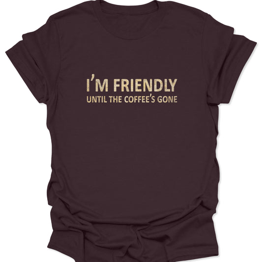 A dark chocolate brown t-shirt with the minimalist quote "I'M FRIENDLY UNTIL THE COFFEE'S GONE" printed in a cream-colored serif and sans-serif font mix.