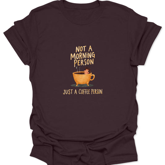 Dark brown t-shirt with a vintage-style coffee graphic: a tiny figure submerged in an overflowing, hot coffee mug. Text reads: "NOT A MORNING PERSON, JUST A COFFEE PERSON."