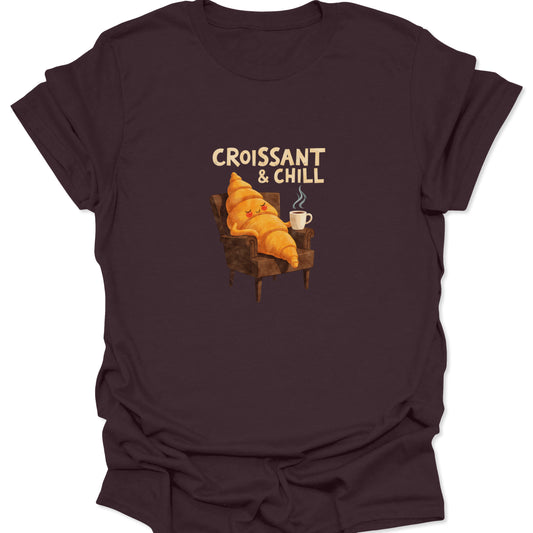 Vintage-style 'Croissant & Chill' graphic tee. Anthropomorphic croissant character relaxing in an easy chair with a hot drink on a deep dark chocolate brown shirt.