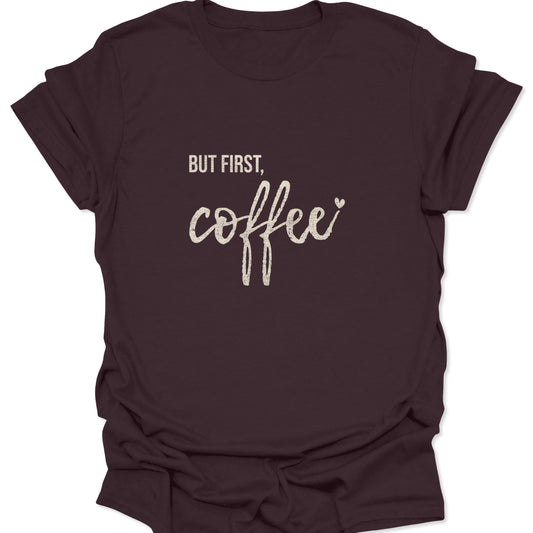 A dark chocolate brown t-shirt with a vintage white distressed print reading "BUT FIRST, coffee" in a mix of sans-serif and cursive fonts.