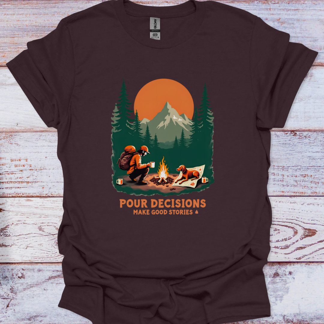 Brown t-shirt with a graphic of a man and his dog by a campfire and mountains, with text 'Pour Decisions Make Good Stories' on a wooden surface.