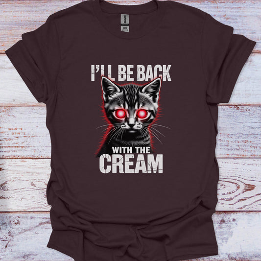 Brown t-shirt with a graphic of a cat with red eyes and text 'I'll be back with the cream' on a wooden surface.