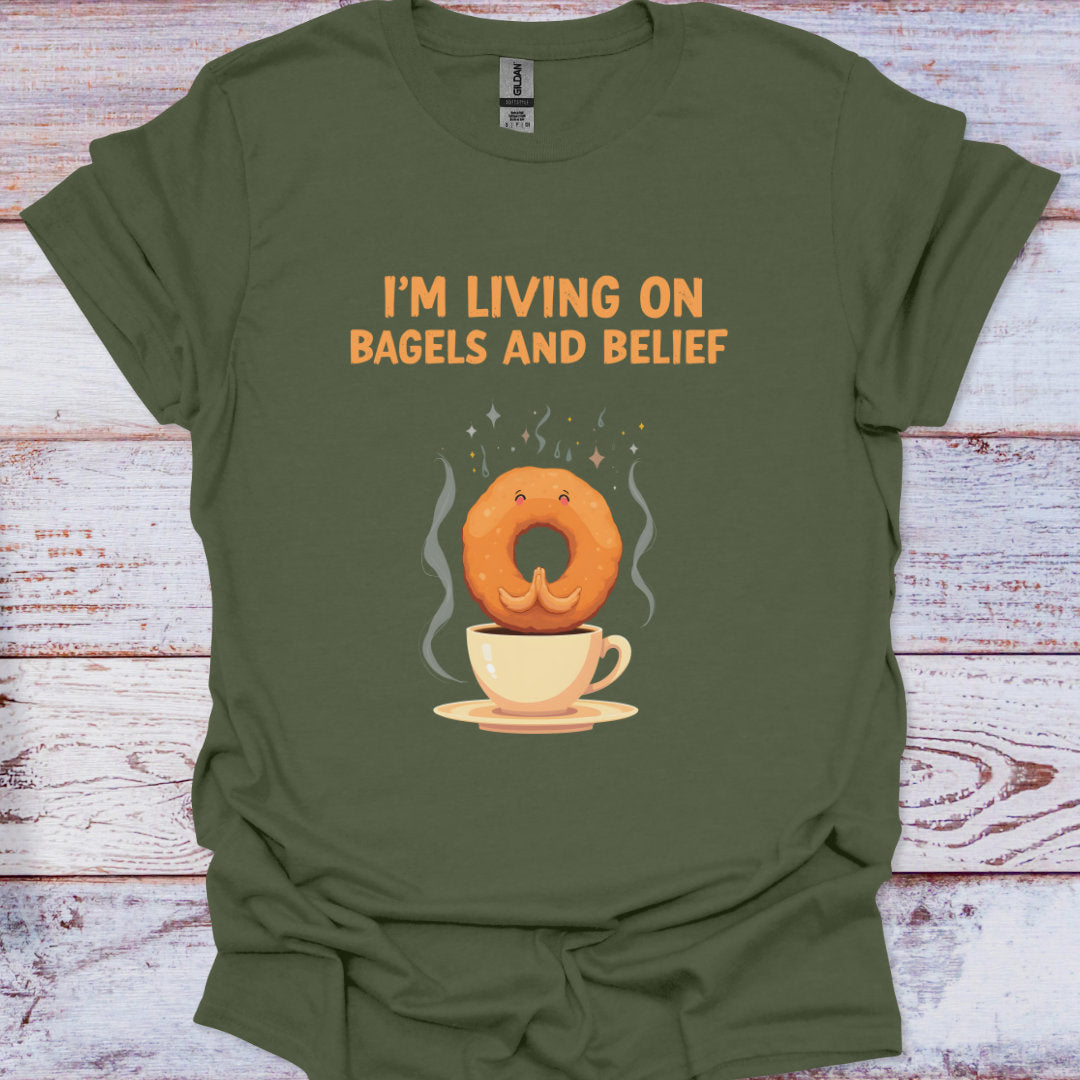 Green t-shirt with a graphic of a bagel in a coffee cup and text 'I'm living on bagels and belief' on a wooden surface.