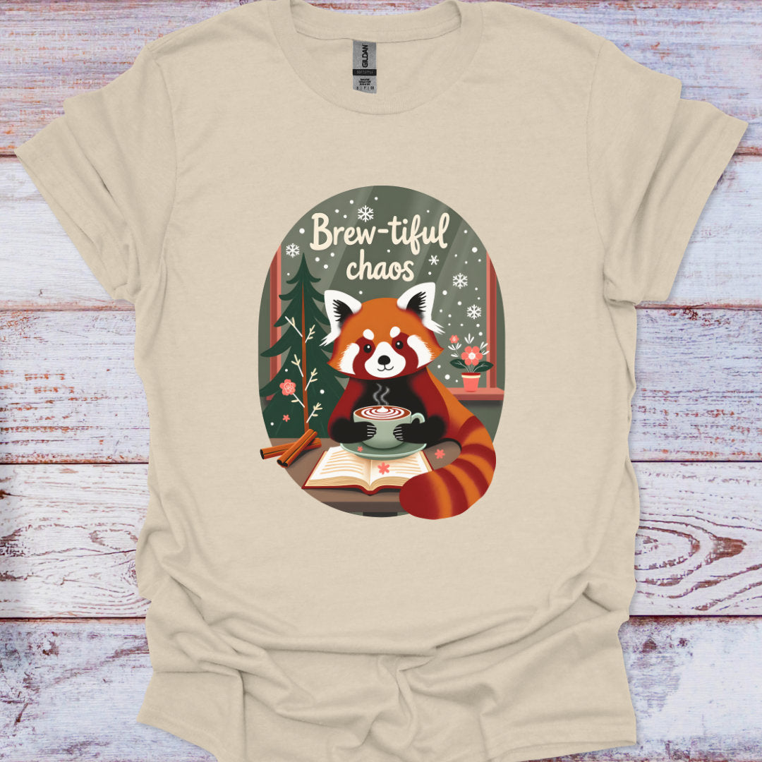 Beige t-shirt with a red panda holding a cup of coffee and the text 'Brew-tiful chaos' on a wooden surface.