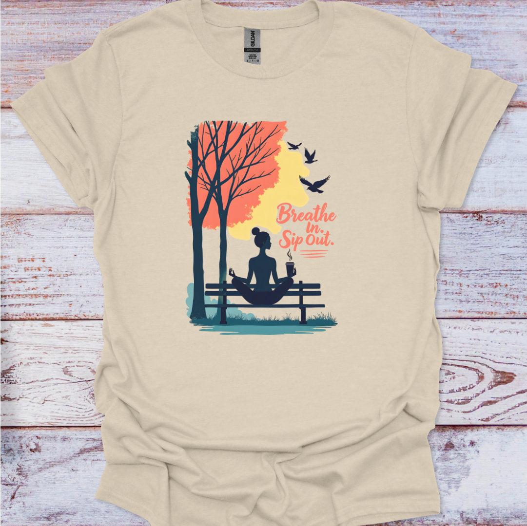 Beige t-shirt with a graphic of a woman meditating by a tree, birds, and text 'Breathe In Sip Out' on a wooden surface.