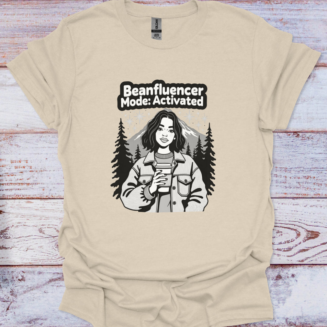 Beige t-shirt with graphic design of a person holding a coffee cup and text 'Beanfluencer Mode: Activated' on a wooden surface.