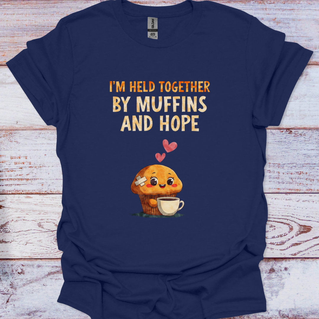 Navy blue t-shirt with a cartoon muffin holding a cup and text 'I'm held together by muffins and hope' on a wooden surface.
