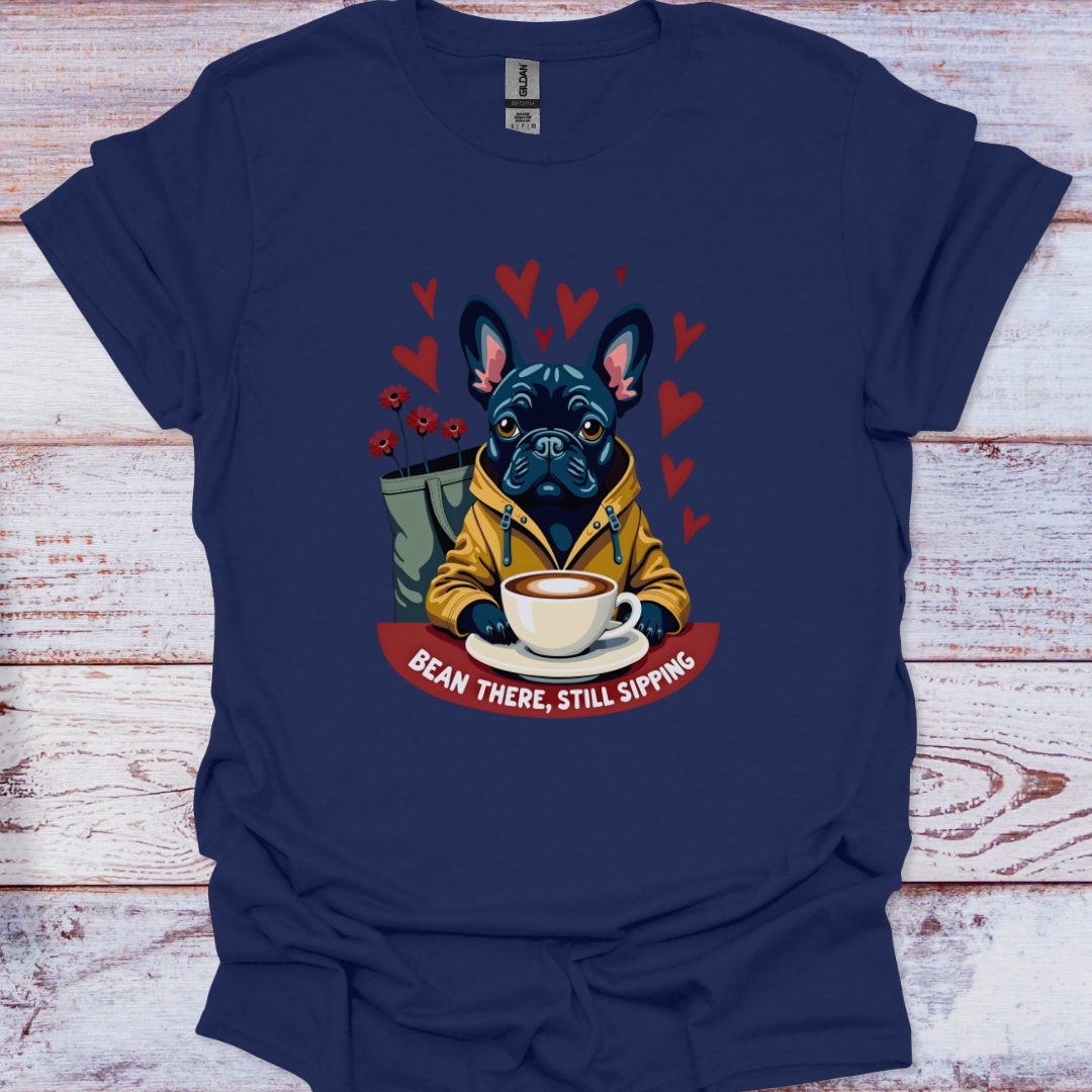 Blue t-shirt with a graphic of a dog holding a coffee cup and text 'Bean There, Still Sipping' on a wooden surface.