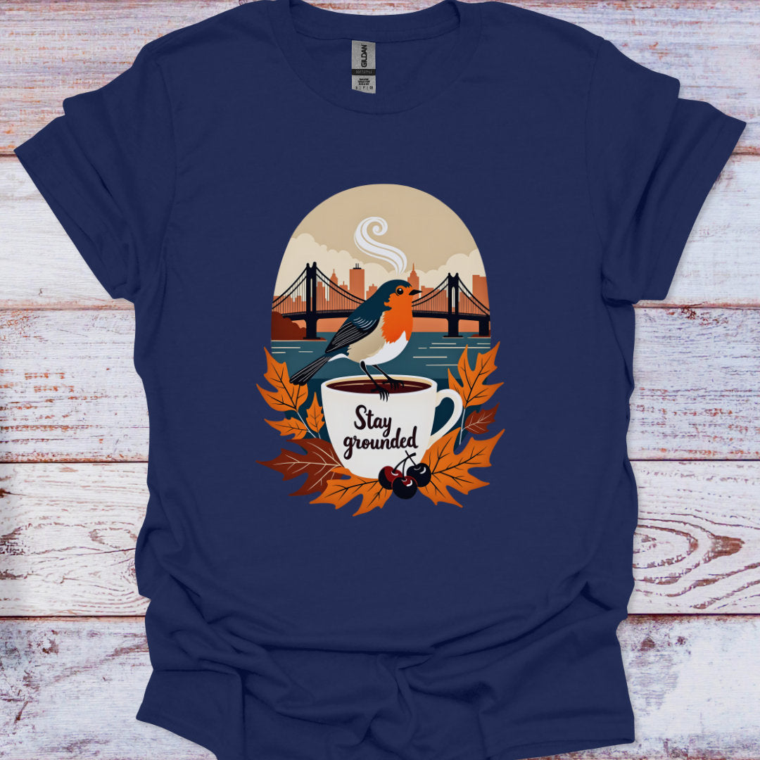 Navy blue t-shirt with a graphic of a bird on a cup with leaves and text 'Stay grounded' on a wooden surface.