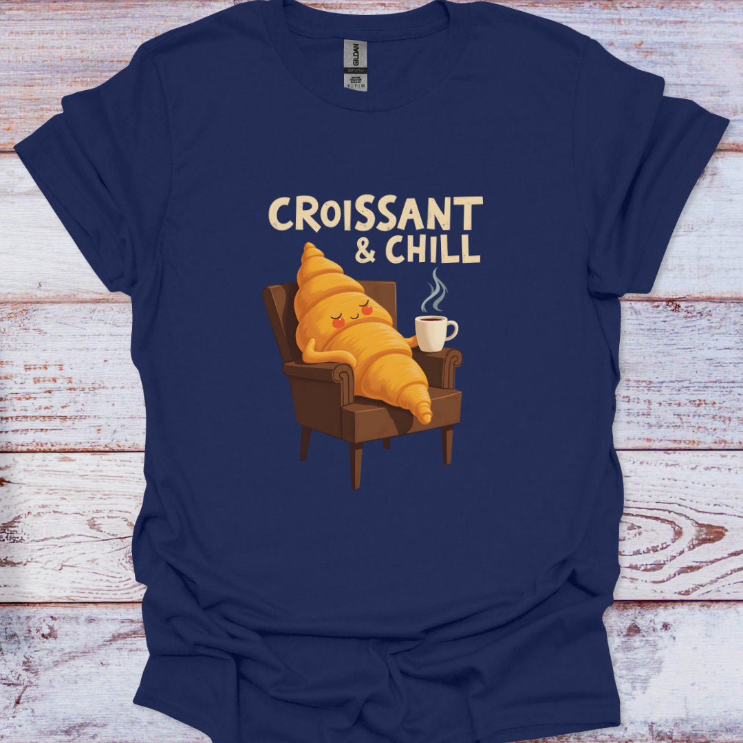 Navy blue t-shirt with a cartoon croissant character sitting in an armchair with a cup of coffee, text 'Croissant & Chill' on a wooden surface.
