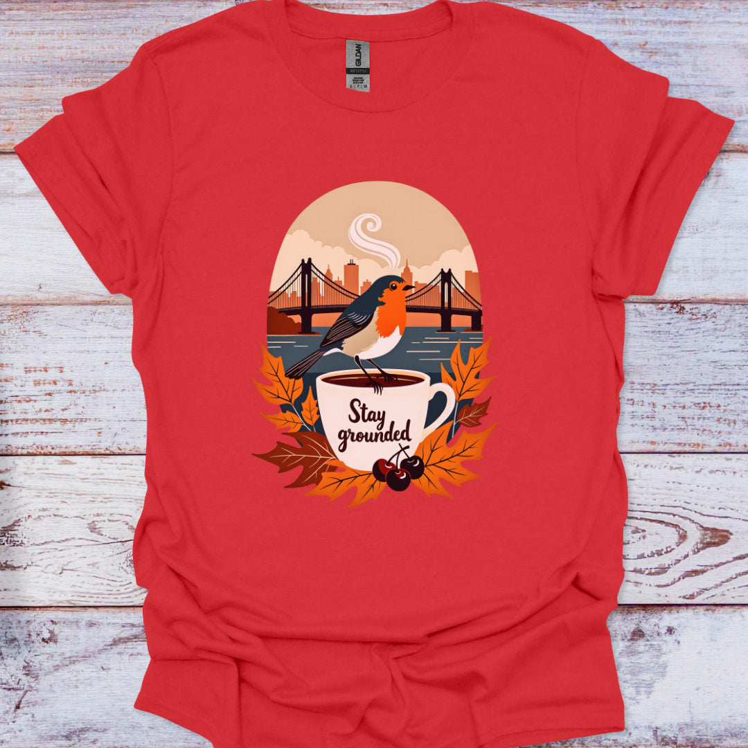 Red t-shirt with a graphic of a bird on a cup with 'Stay grounded' text, set against a wooden background.