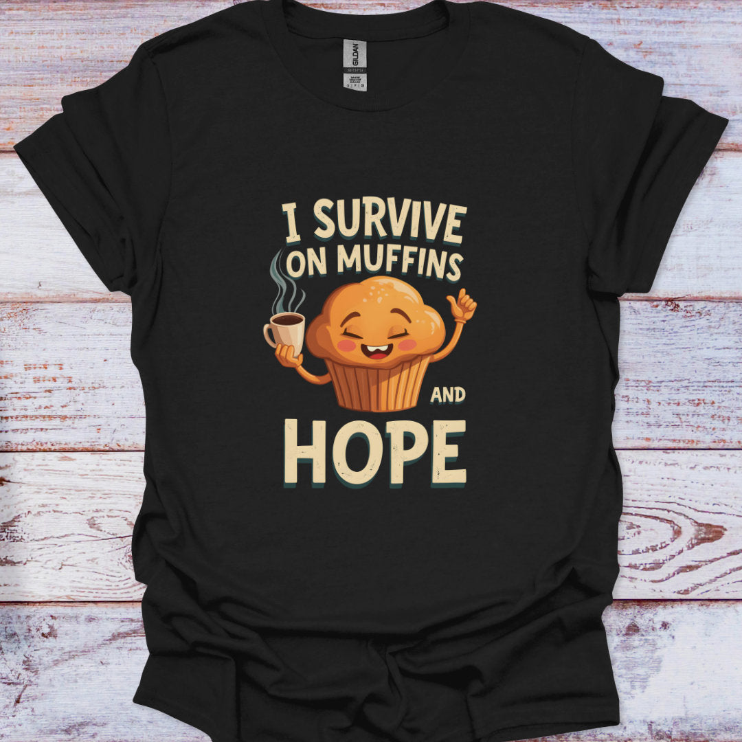 Black t-shirt with a graphic of a muffin holding a coffee cup and text 'I survive on muffins and hope' on a wooden surface.