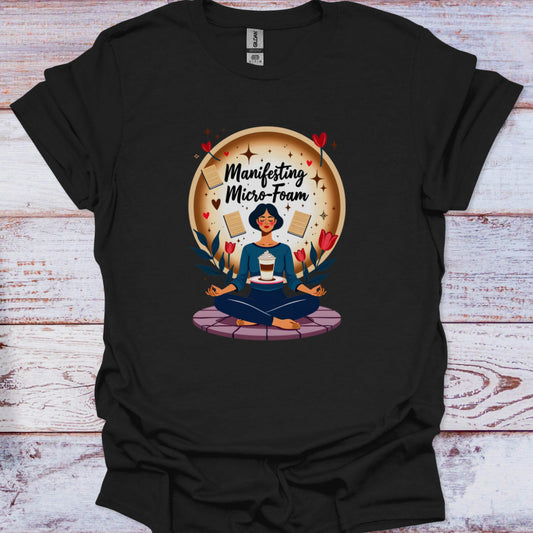 Black t-shirt with a graphic of a person meditating and text 'Manifesting Micro-Foam' on a wooden surface.