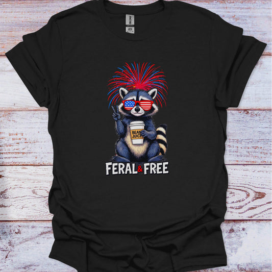 Black t-shirt with a raccoon graphic holding a coffee cup, wearing sunglasses, and text 'Feral & Free' on a wooden surface.