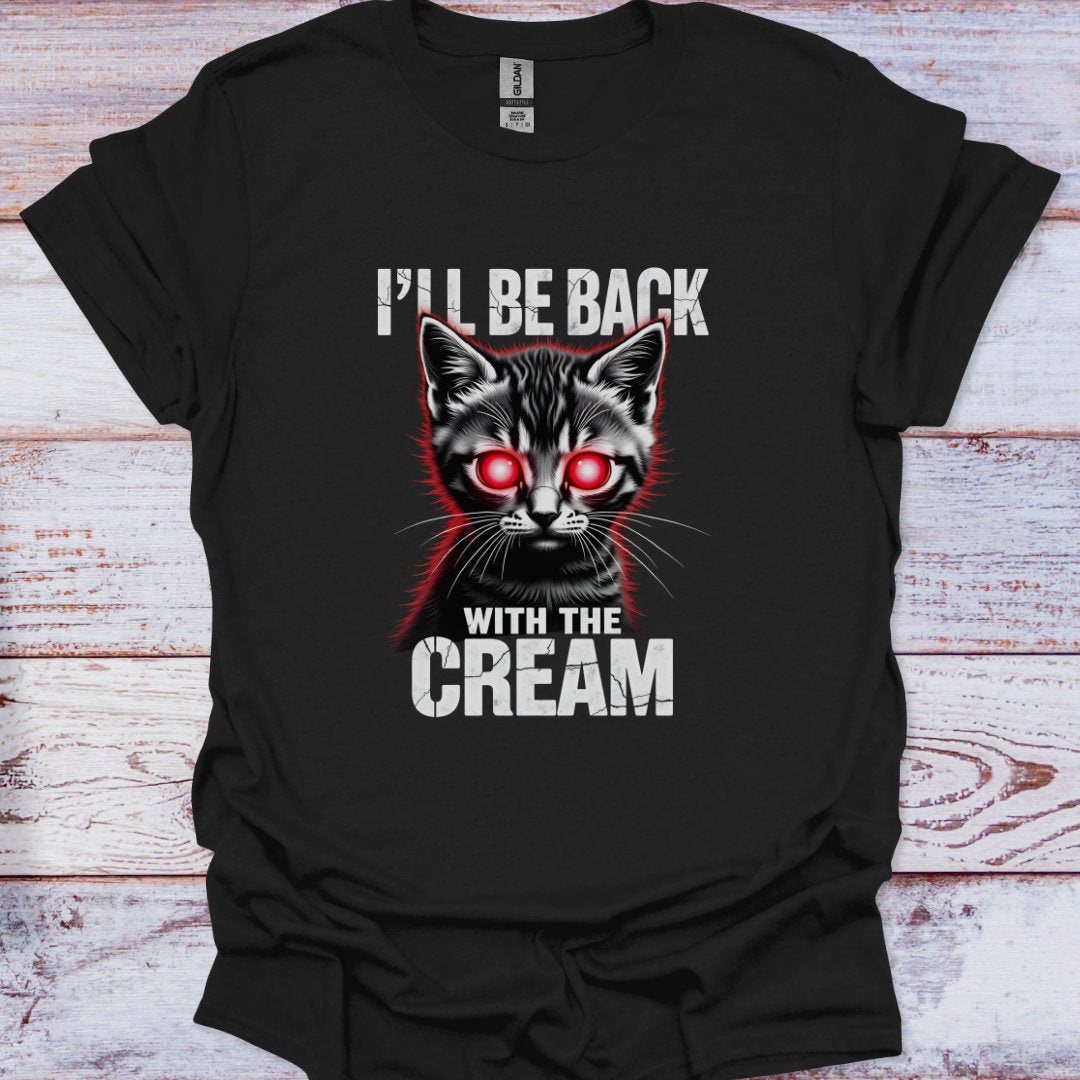 Black t-shirt with a graphic of a cat with red eyes and text 'I'll be back with the cream' on a wooden surface.