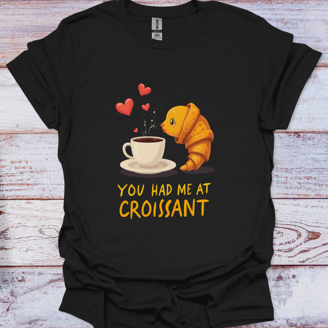 Black t-shirt with a croissant and coffee cup graphic and text 'You Had Me at Croissant' on a wooden surface.
