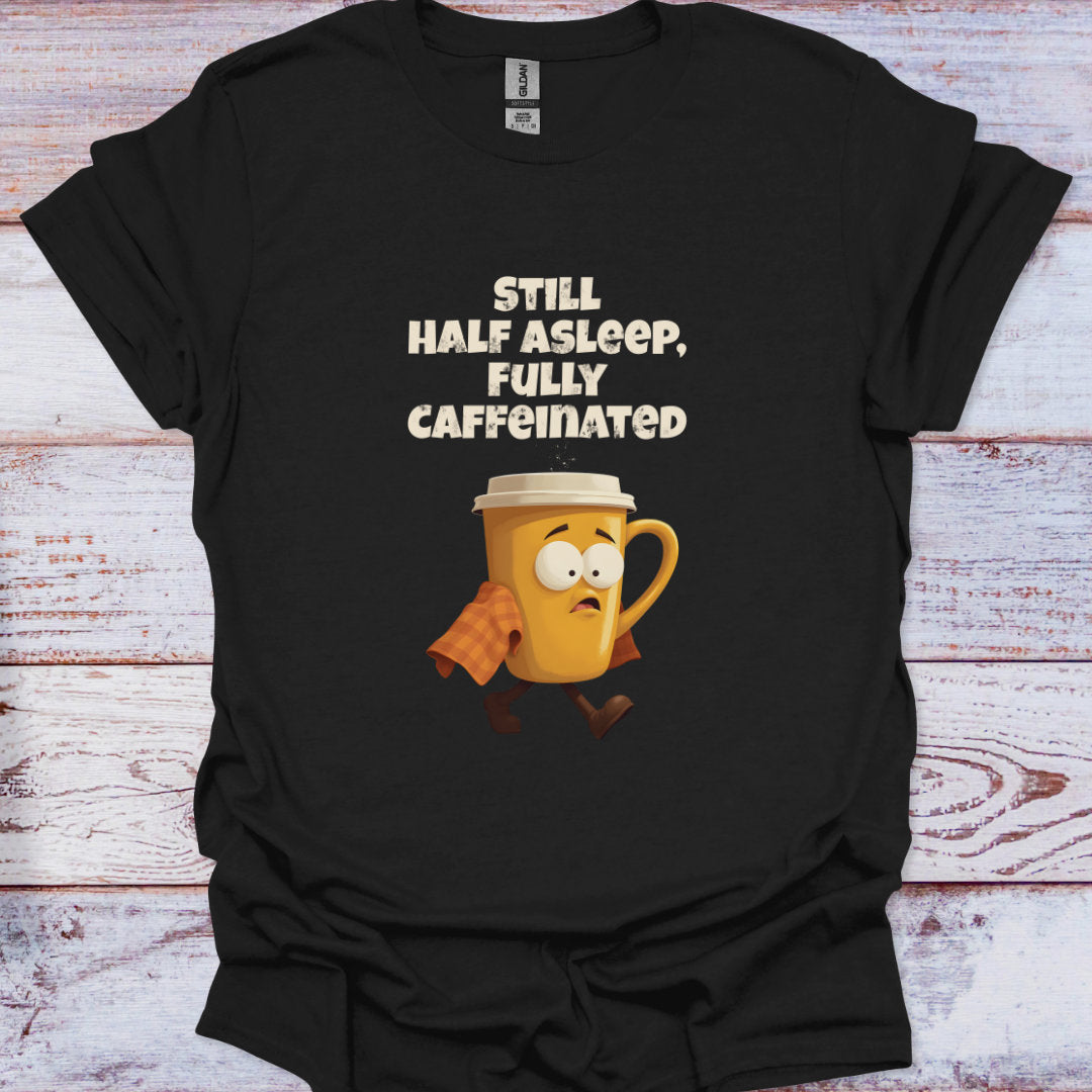 Black t-shirt with a cartoon coffee mug and text 'Still half asleep, fully caffeinated' on a wooden surface.