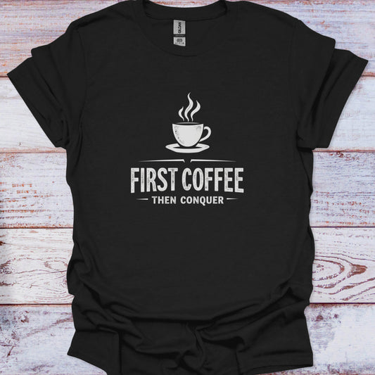 Black t-shirt with a coffee cup graphic and text 'First Coffee Then Conquer' on a wooden surface.