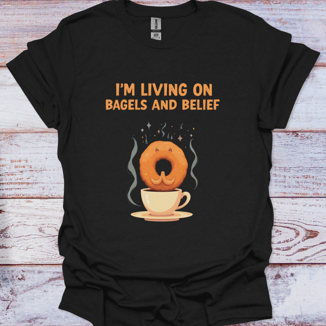 Black t-shirt with a graphic of a bagel and coffee cup and text 'I'm living on bagels and belief' on a wooden surface.