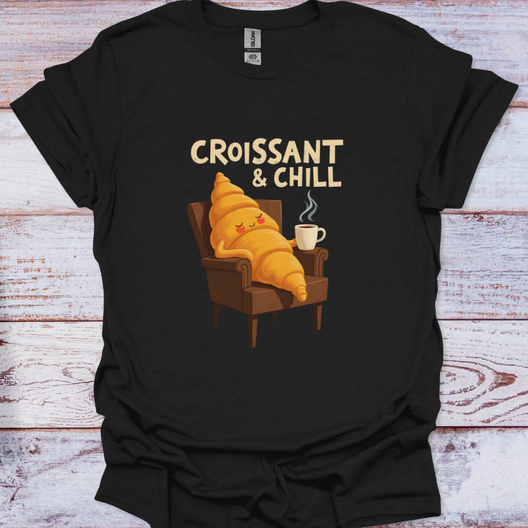 Black t-shirt with a cartoon croissant character sitting on an armchair with a cup of coffee, text 'Croissant & Chill' on a wooden surface.