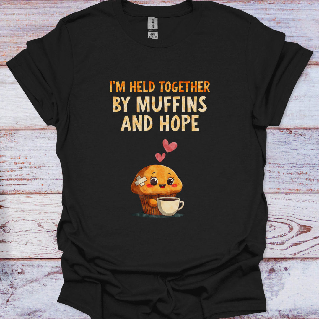Black t-shirt with a cartoon muffin and text 'I'm held together by muffins and hope' on a wooden surface
