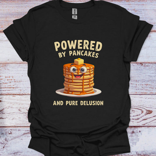 Black t-shirt with a graphic of pancakes and text 'Powered by Pancakes and Pure Delusion' on a wooden surface.