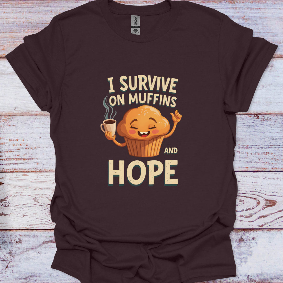 Brown t-shirt with a cartoon muffin holding a coffee cup and text 'I Survive on Muffins and Hope' on a wooden surface.