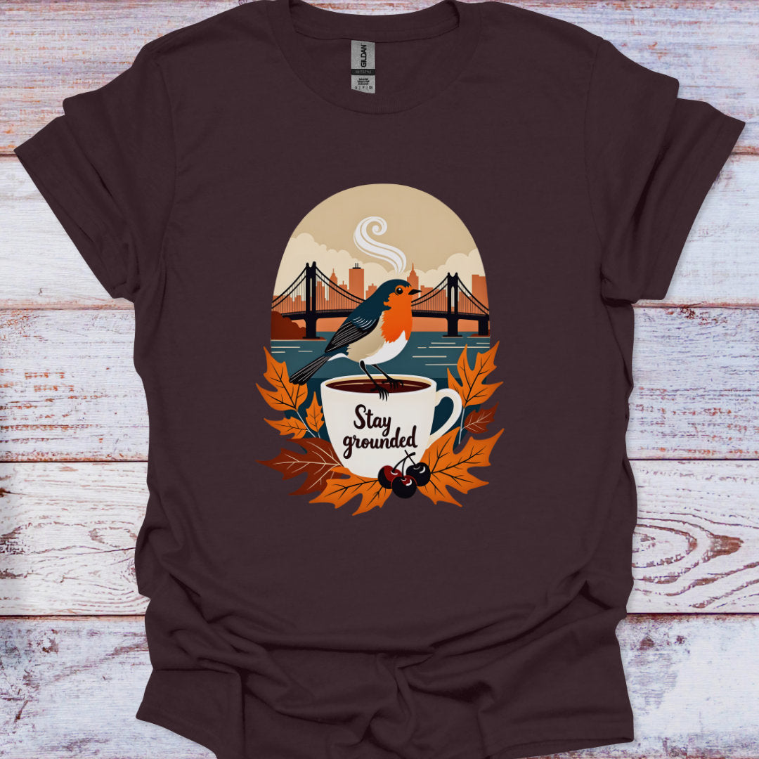 Brown t-shirt with a graphic of a bird on a coffee cup and 'Stay grounded' text, placed on a wooden surface.