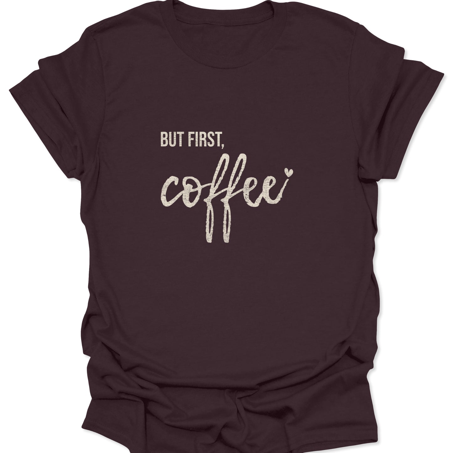 A dark chocolate brown t-shirt with a vintage white distressed print reading "BUT FIRST, coffee" in a mix of sans-serif and cursive fonts.
