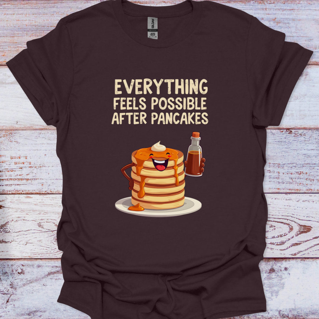 Brown t-shirt with a graphic of pancakes and syrup with text 'Everything feels possible after pancakes' on a wooden surface.