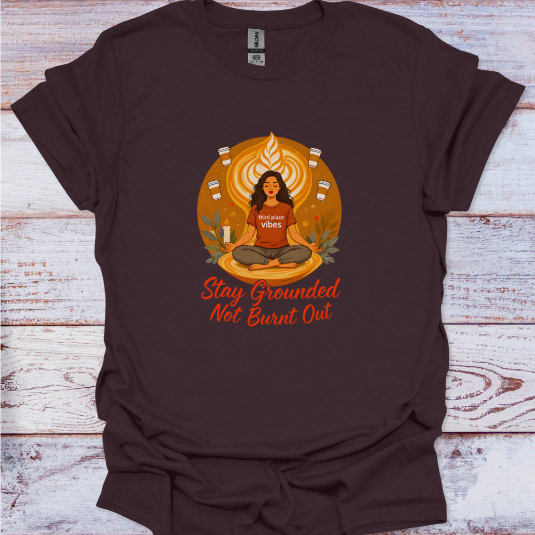 Brown t-shirt with a graphic of a person in meditation and text 'Stay Grounded Not Burnt Out' on a wooden surface.
