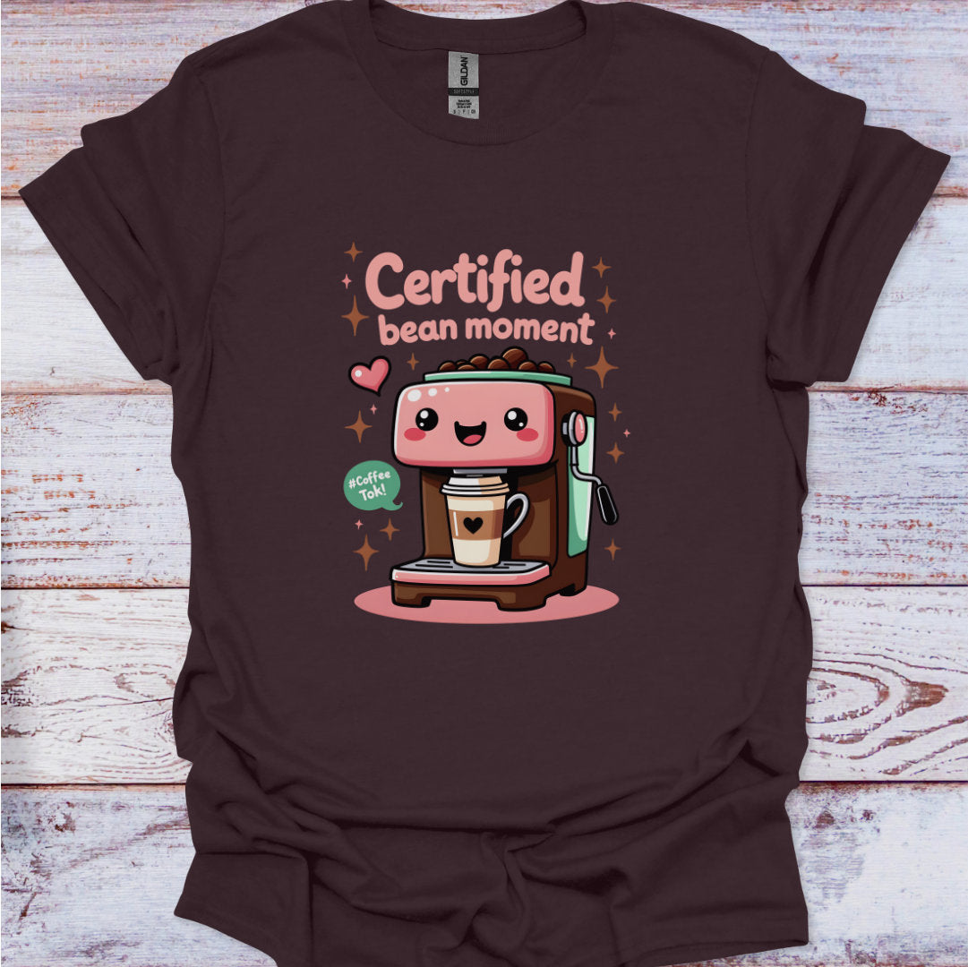 Brown t-shirt with a cartoon coffee machine and text 'Certified bean moment' on a wooden surface.