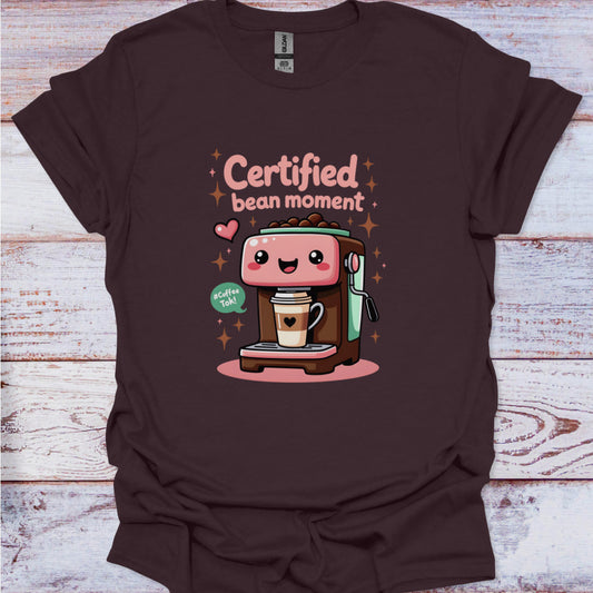 Brown t-shirt with a cartoon coffee machine and text 'Certified bean moment' on a wooden surface.