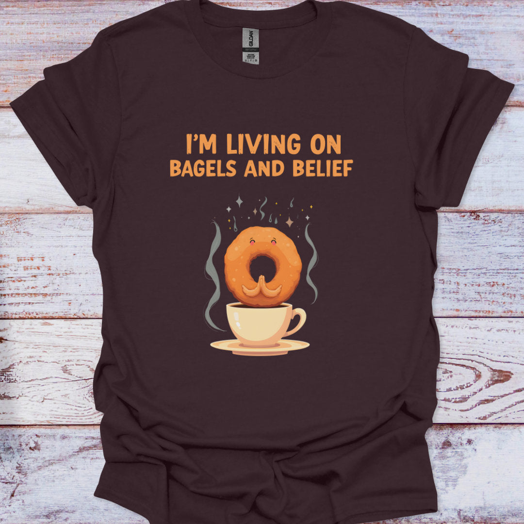 Brown t-shirt with a graphic of a bagel and coffee cup and text 'I'm living on bagels and belief' on a wooden surface.