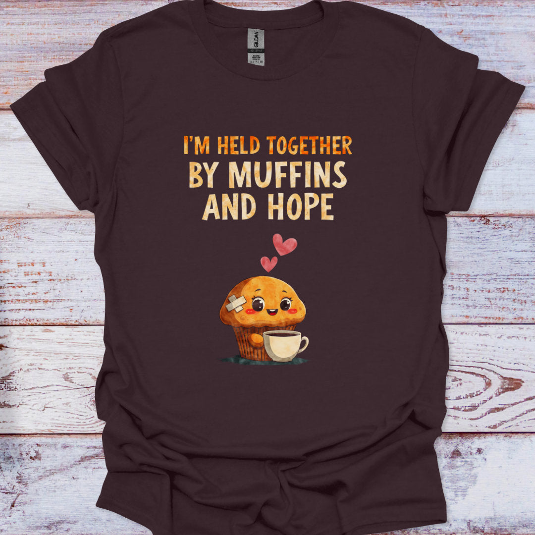 Brown t-shirt with a cartoon muffin and text 'I'm held together by muffins and hope' on a wooden surface.