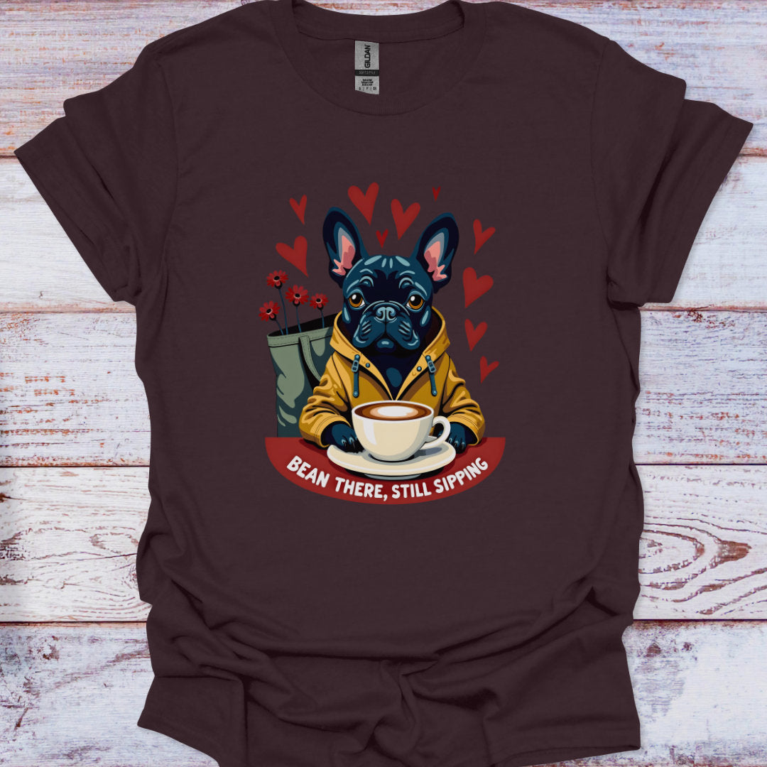 Brown t-shirt with a graphic of a dog holding a coffee cup and text 'Bean There, Still Sipping' on a wooden surface.