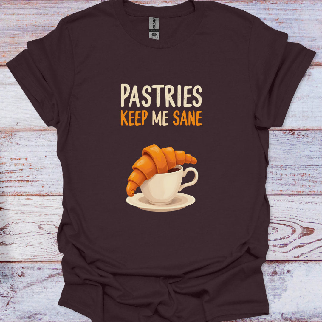 Brown t-shirt with a graphic of a croissant and coffee cup and text 'Pastries Keep Me Sane' on a wooden surface.
