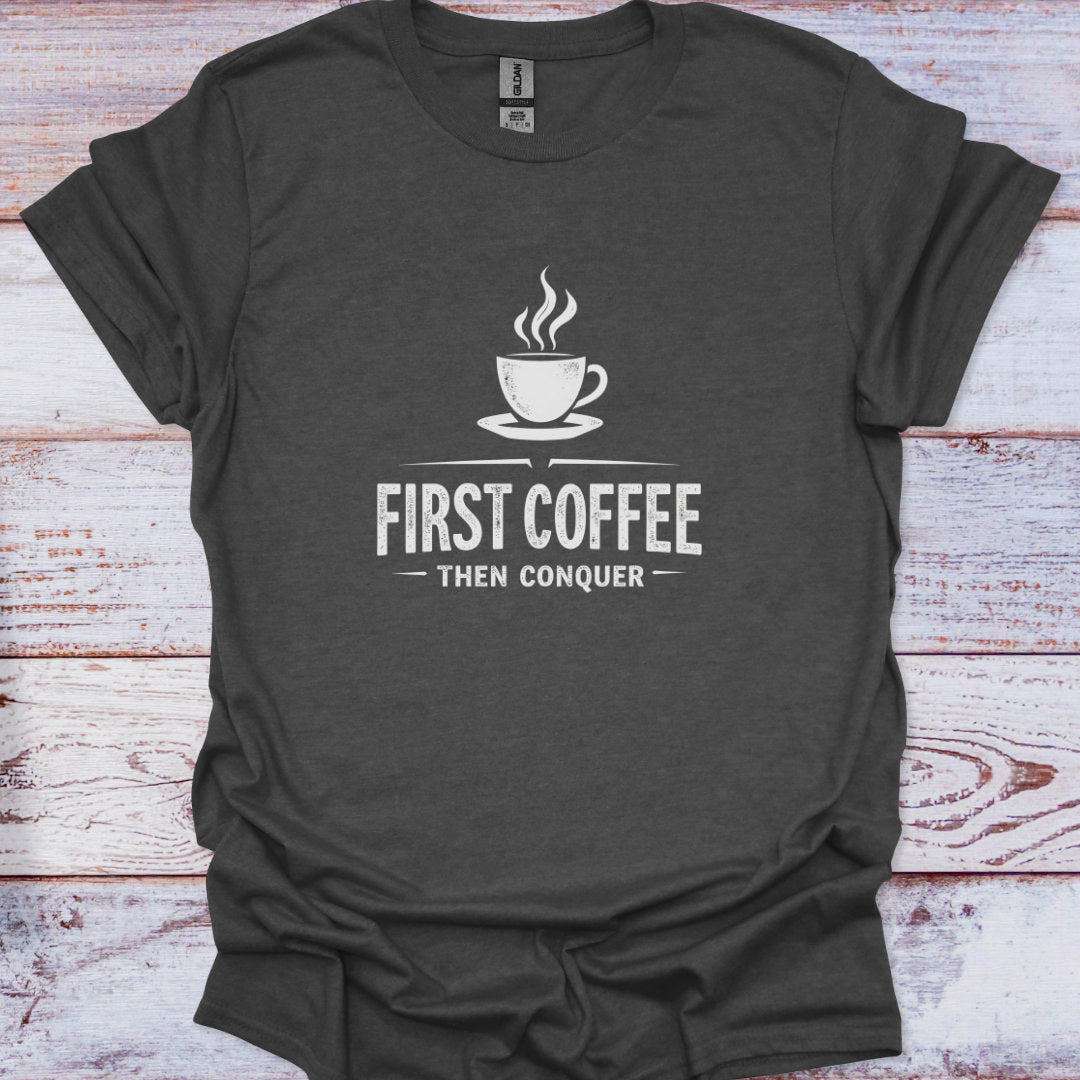 Dark gray t-shirt with 'First Coffee Then Conquer' text and coffee cup graphic on a wooden surface
