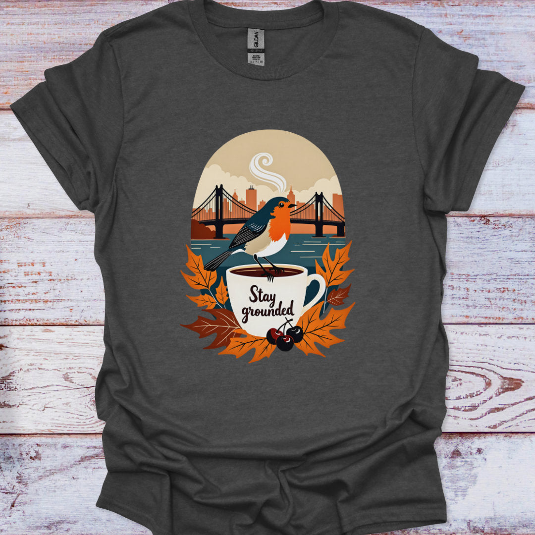 Dark gray t-shirt with a graphic of a bird on a coffee cup and 'Stay grounded' text, placed on a wooden surface.