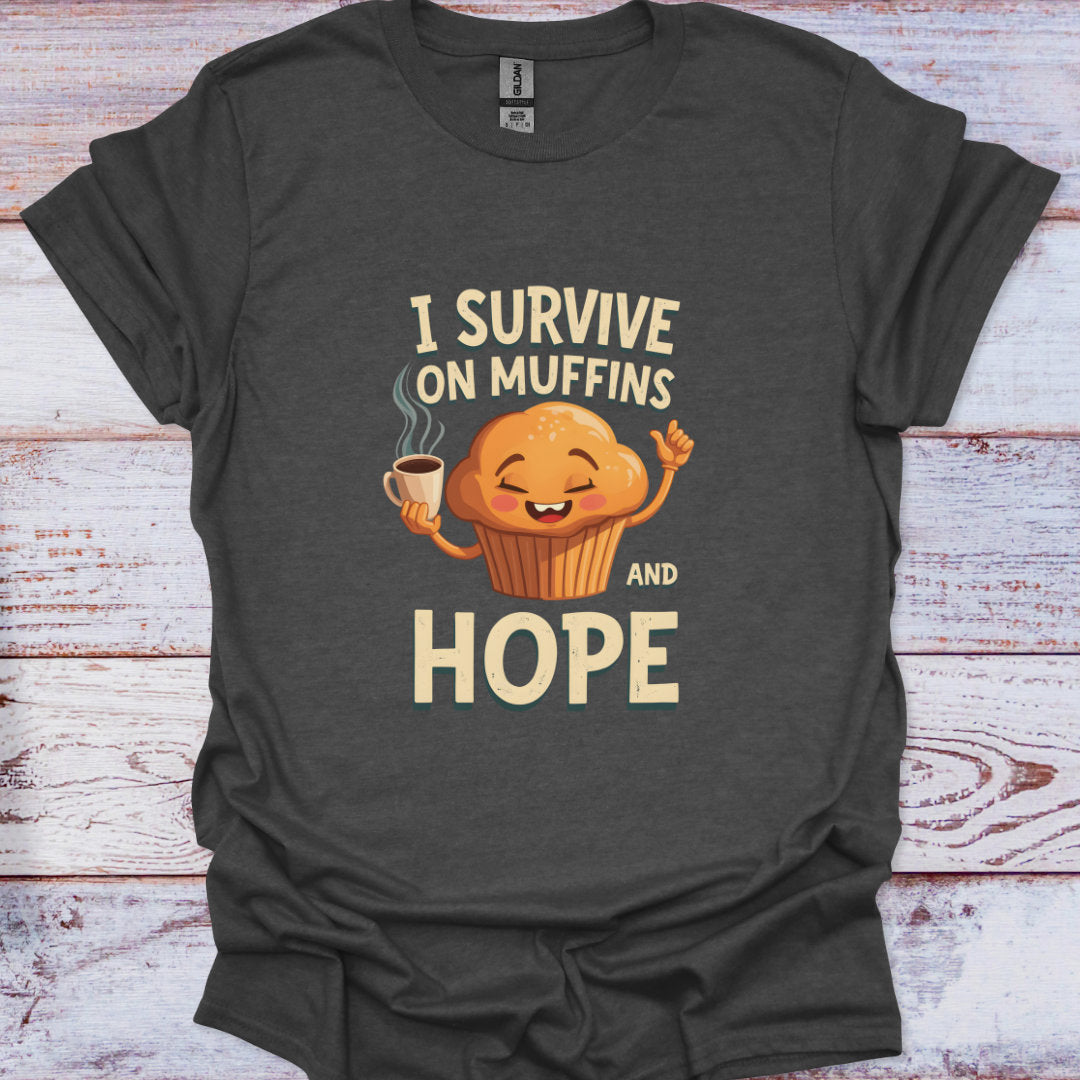 Dark gray t-shirt with a cartoon muffin holding a coffee cup and text 'I Survive on Muffins and Hope' on a wooden surface.