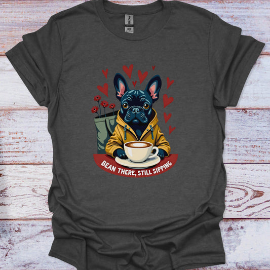 Grey t-shirt with a graphic of a dog holding a coffee cup and text 'Bean There, Still Sipping' on a wooden surface.