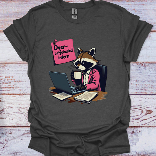 Gray t-shirt with a raccoon illustration holding a coffee cup and using a laptop, on a wooden surface.