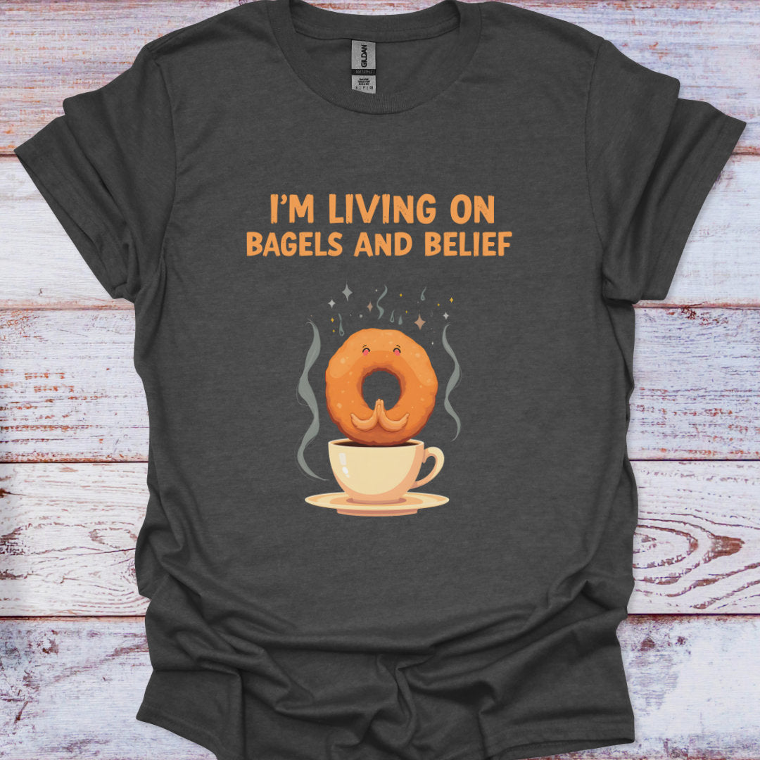 Dark gray t-shirt with a graphic of a bagel on a cup of coffee and text 'I'm living on bagels and belief' on a wooden surface.