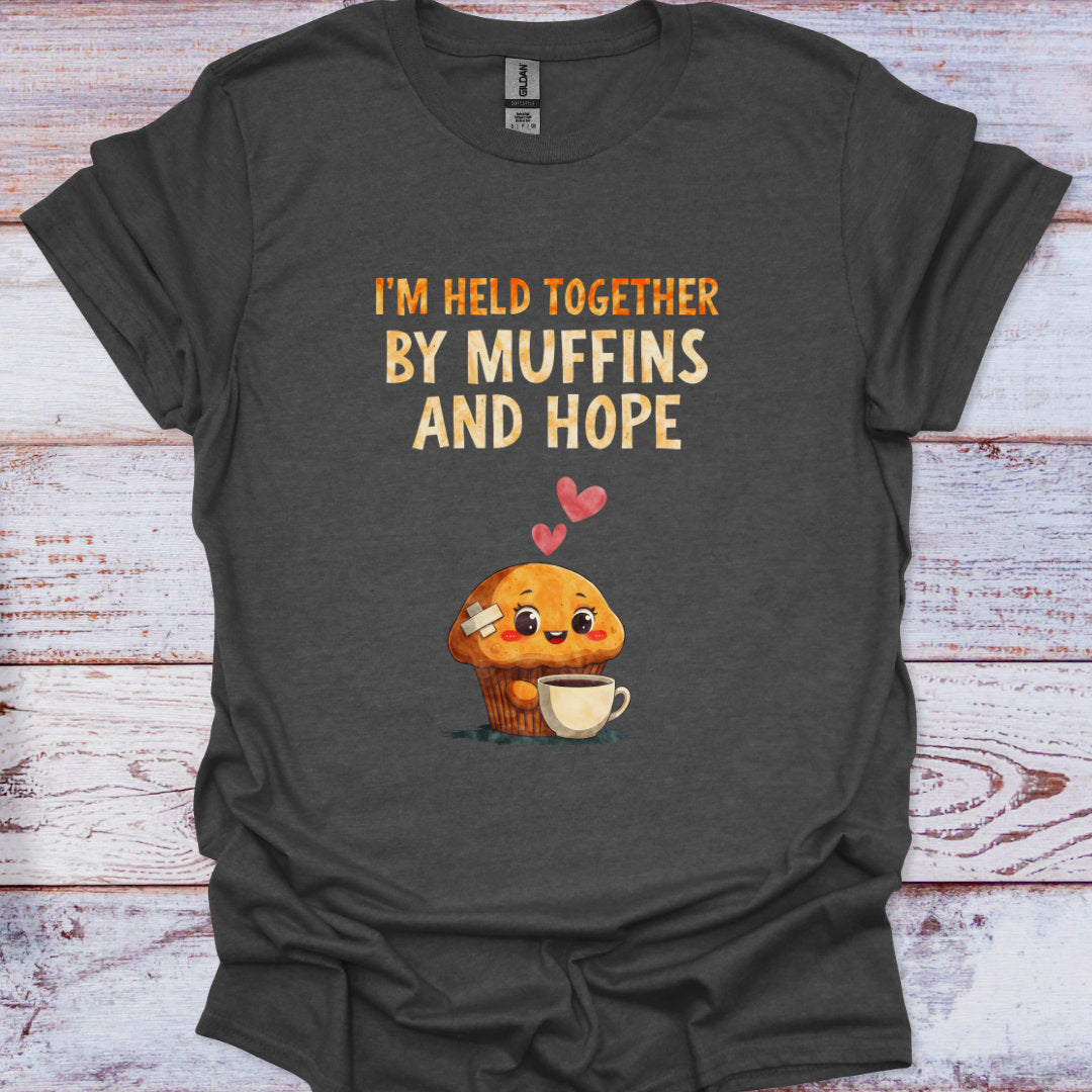 Dark gray t-shirt with a cartoon muffin holding a cup and text 'I'm held together by muffins and hope' on a wooden surface.
