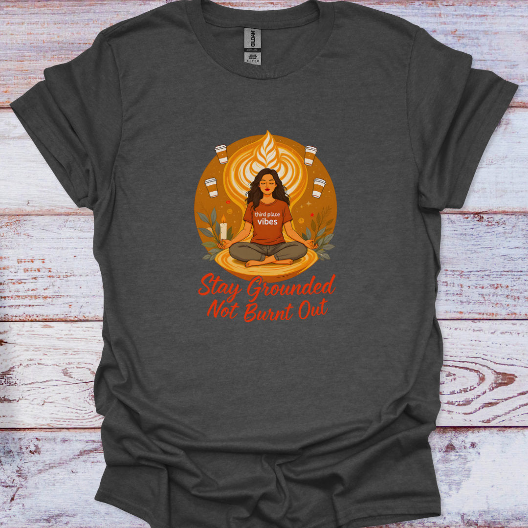 Dark gray t-shirt with a graphic of a person meditating and text 'Stay Grounded Not Burnt Out' on a wooden surface.