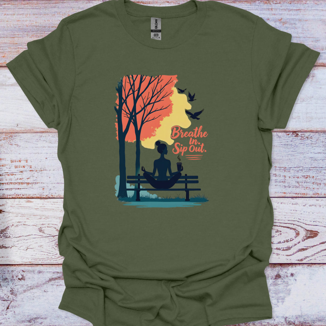 Green t-shirt with a graphic of a person meditating by a tree, birds, and sunset text 'Breathe In Sip Out' on a wooden surface.