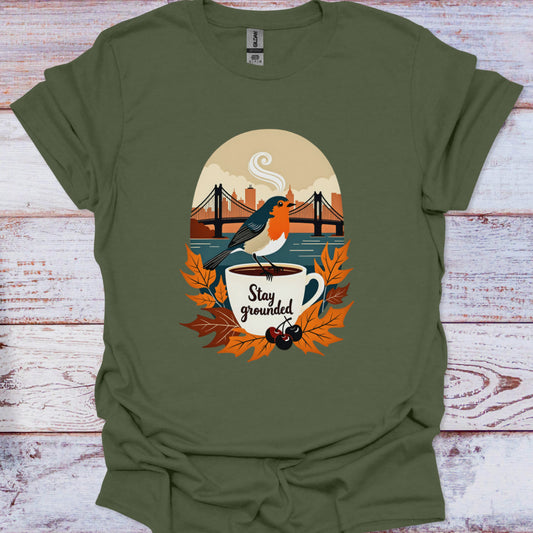 Green t-shirt with a graphic of a bird on a coffee cup and 'Stay grounded' text, placed on a wooden surface.
