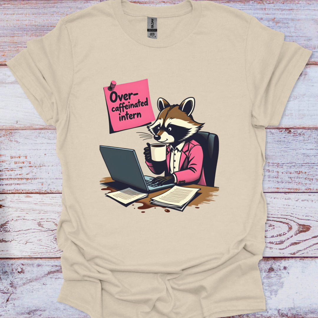 Beige t-shirt with a raccoon graphic holding a coffee cup and using a laptop, with a pink sign reading 'Overcaffeinated intern' on a wooden surface.