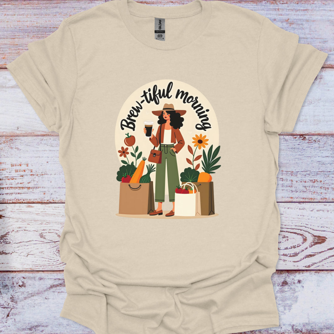 Beige t-shirt with a graphic of a woman holding a coffee cup and shopping bags, with 'Brew-iful morning' text, on a wooden surface.